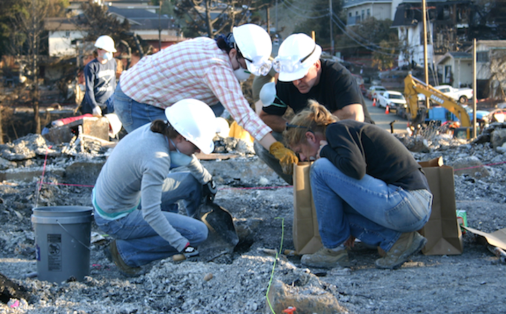 Four people in hard hats bent over and digging for artifacts